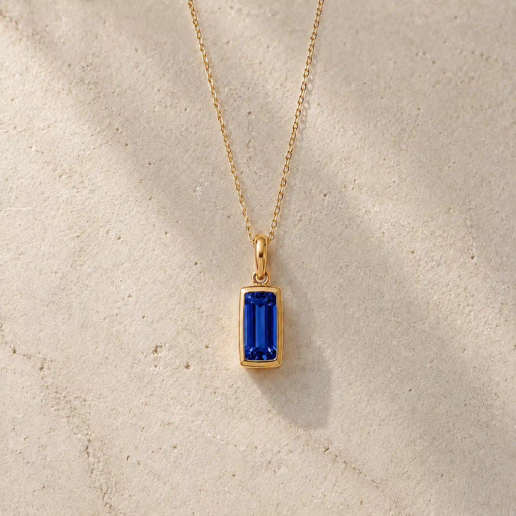 Gold fine chain necklace with a rectangle faceted bezel set september cubic zirconia pendant on a light stone background.