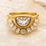 A yellow gold ring featuring a large central diamond in a semi-circular setting, surrounded by smaller baguette and round diamonds, displayed on a light beige stone background.