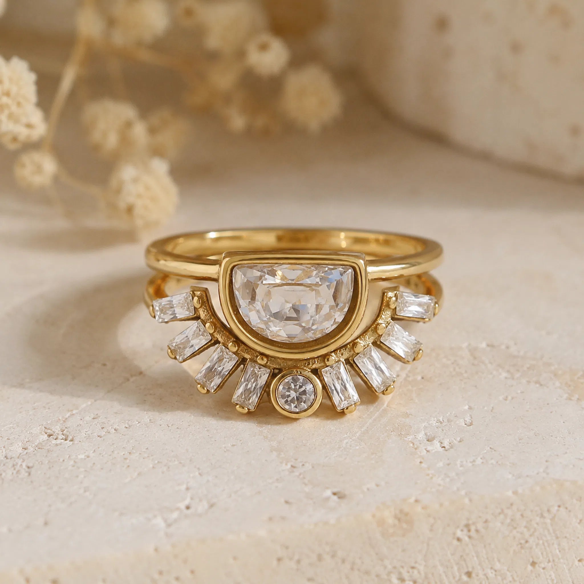 A gold ring with a large clear central gemstone and a fan of smaller clear baguette and round stones, photographed on a light textured surface with soft dried flowers in the background.