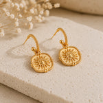 A pair of gold-toned drop earrings with square sunburst medallions, displayed on a textured beige stone slab with dried white flowers in the background.