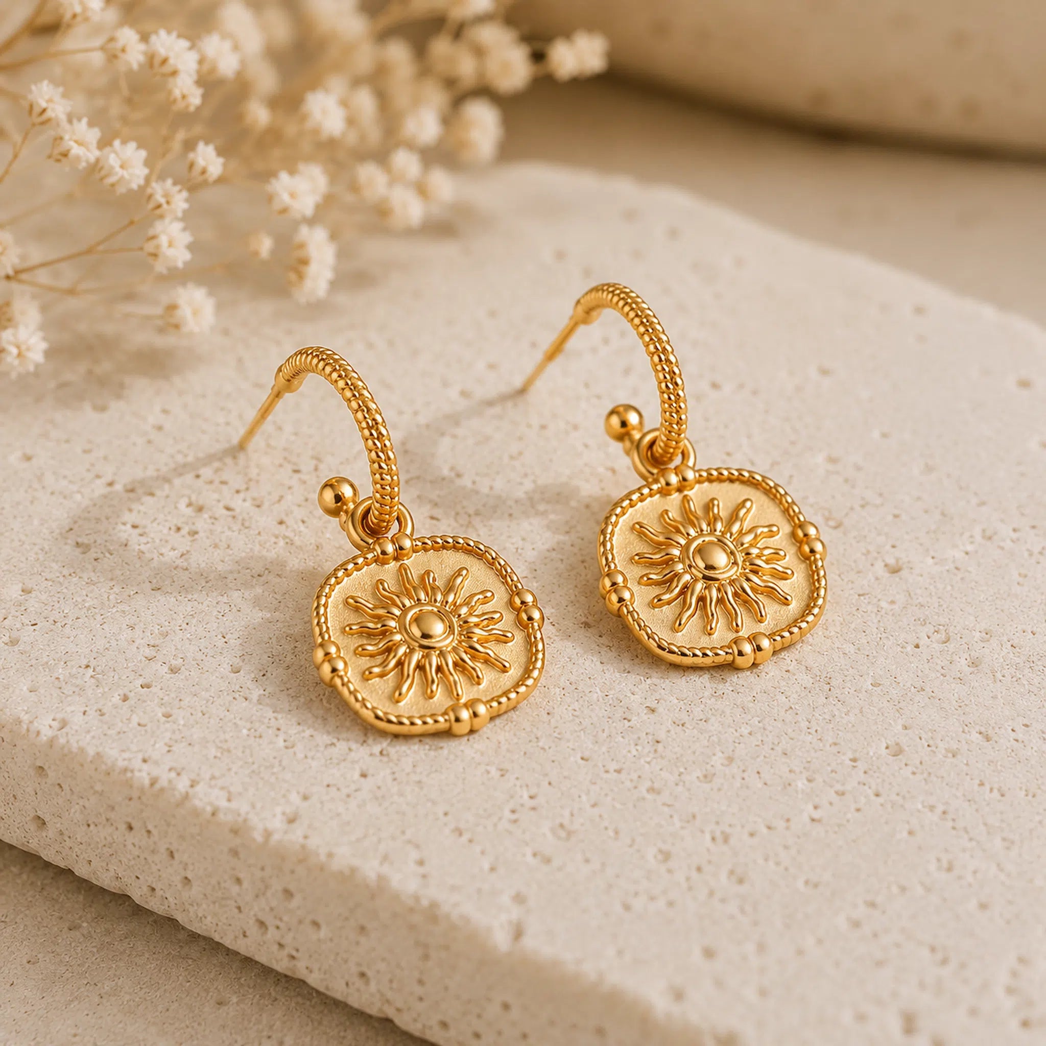 A pair of gold-toned drop earrings with square sunburst medallions, displayed on a textured beige stone slab with dried white flowers in the background.