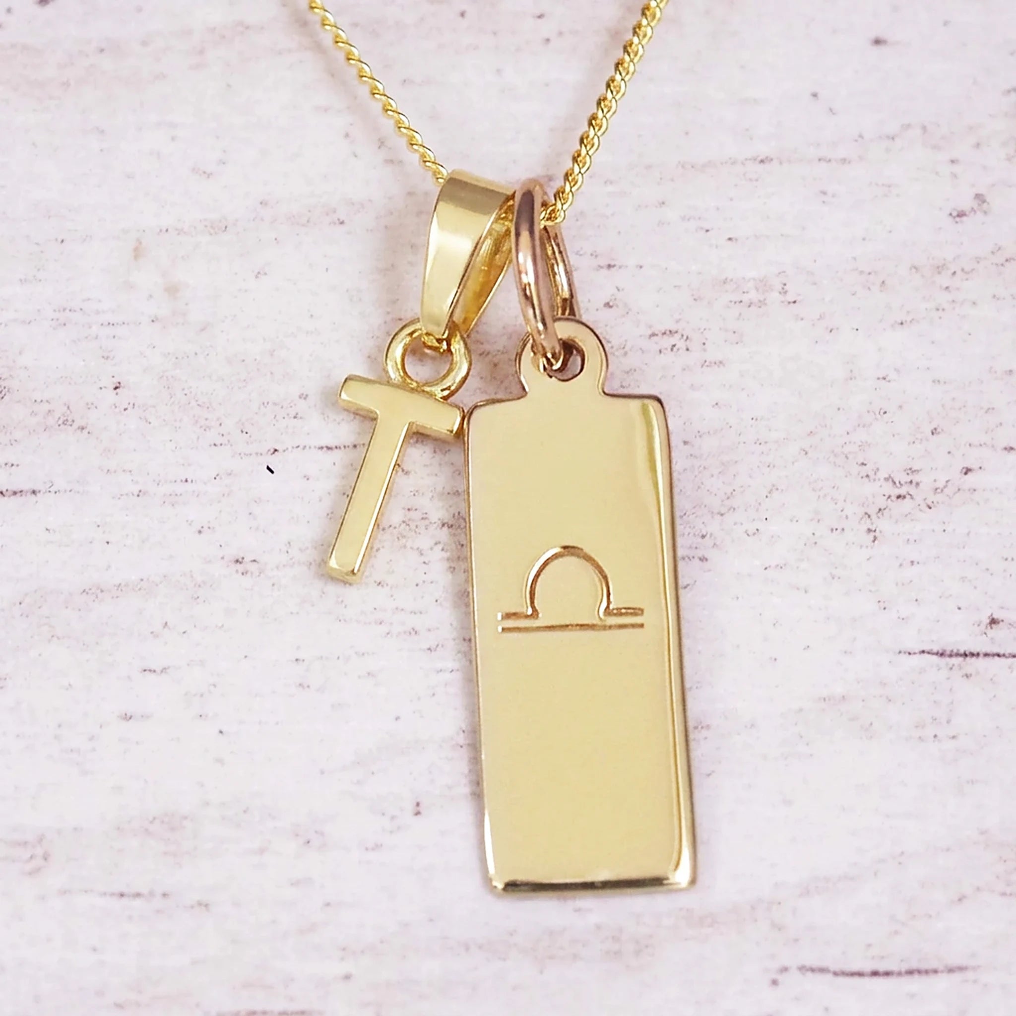 Gold initial and Libra zodiac necklace with a letter T charm and engraved bar pendant on whitewashed wood