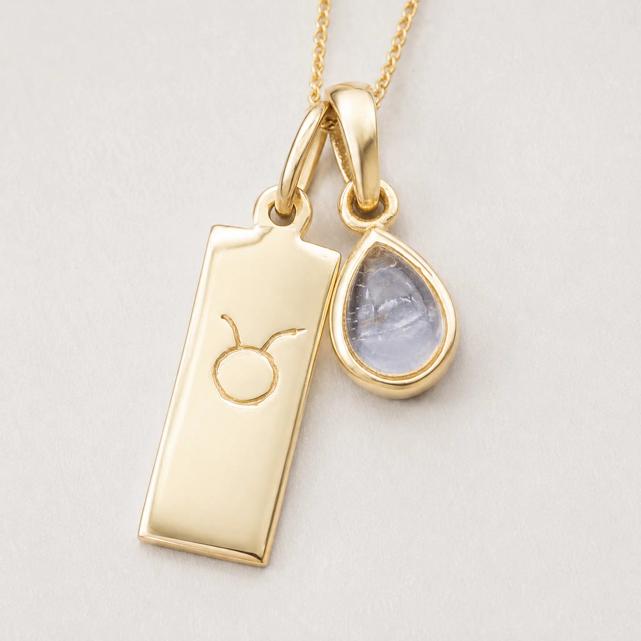 A gold pendant necklace featuring a rectangular bar charm engraved with the taurus zodiac symbol and a teardrop-shaped clear quartz gemstone charm, shown on a light background.
