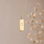 A gold taurus necklace featuring a rectangular pendant necklace engraved with the taurus zodiac symbol, hanging from a delicate gold chain against a warm beige background with blurred dried flowers on the right.