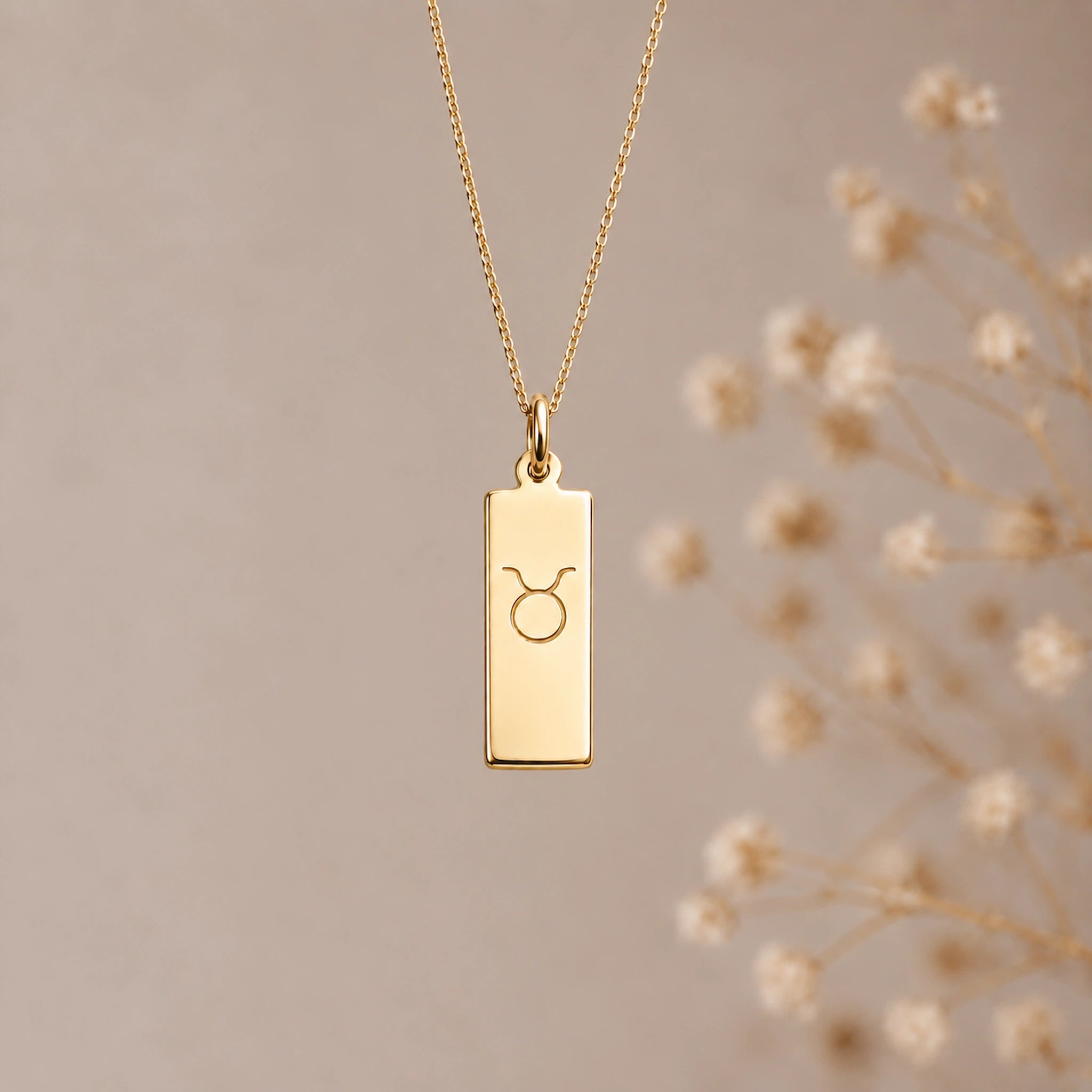 A gold taurus necklace featuring a rectangular pendant necklace engraved with the taurus zodiac symbol, hanging from a delicate gold chain against a warm beige background with blurred dried flowers on the right.