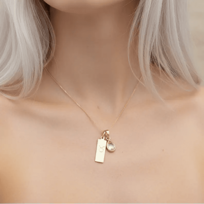 Close-up of a woman wearing a gold necklace with a taurus zodiac pendant and april birthstone pendant.