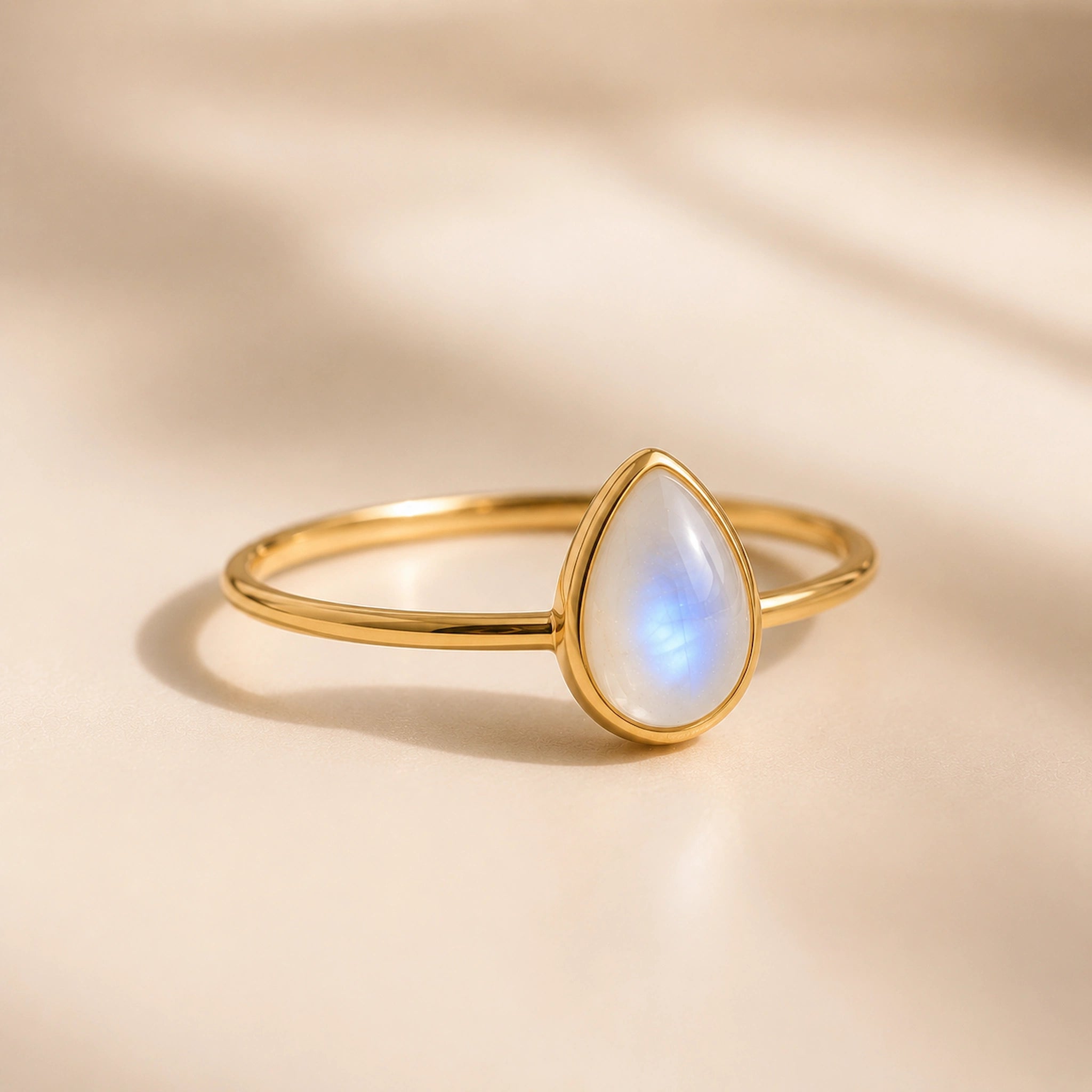 A gold ring with a teardrop-shaped moonstone gemstone, photographed on a warm beige background with soft shadows.