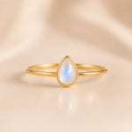A delicate gold ring featuring a teardrop-shaped moonstone gemstone with a soft blue and white iridescent sheen, set against a warm beige satin background.