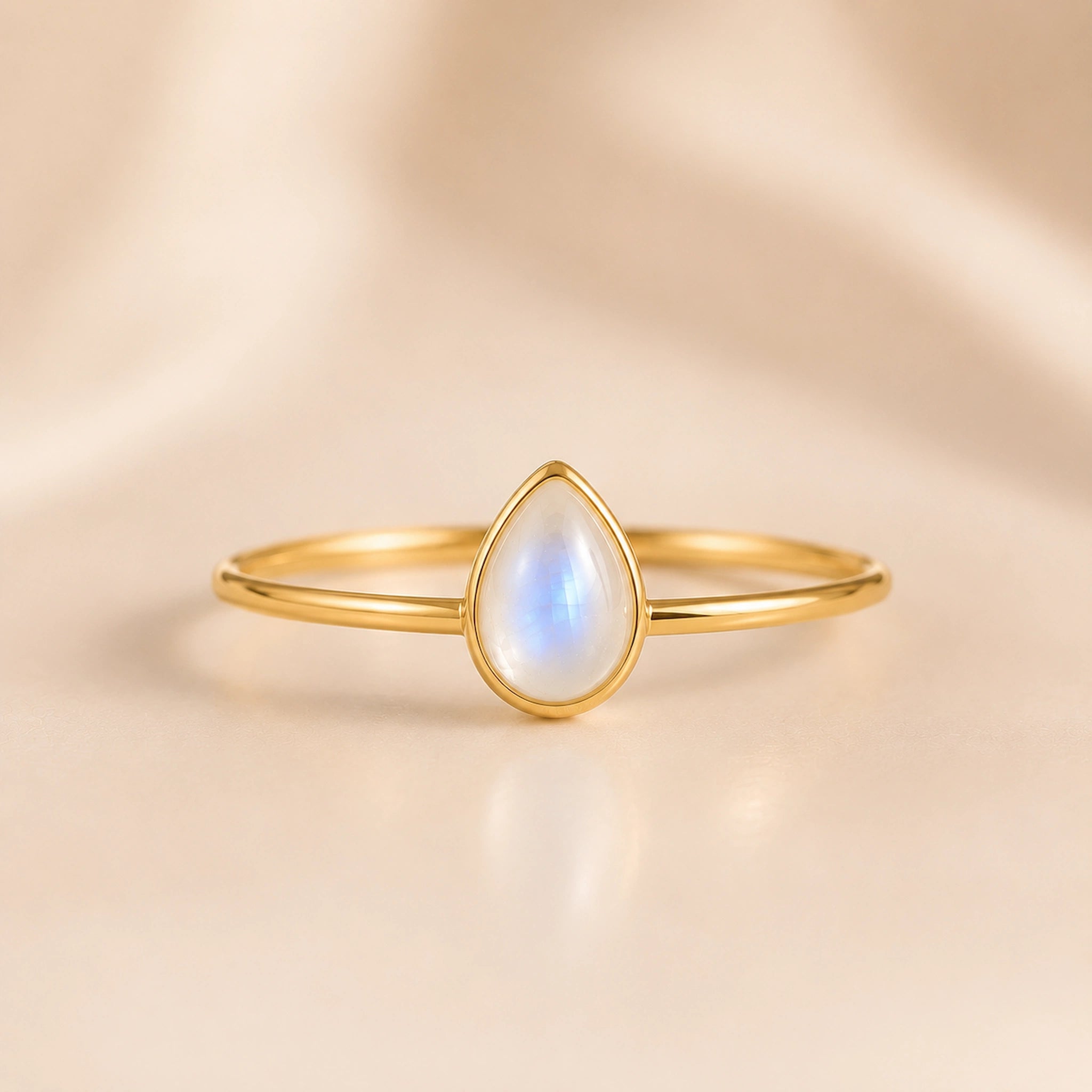 A delicate gold ring featuring a teardrop-shaped moonstone gemstone with a soft blue and white iridescent sheen, set against a warm beige satin background.