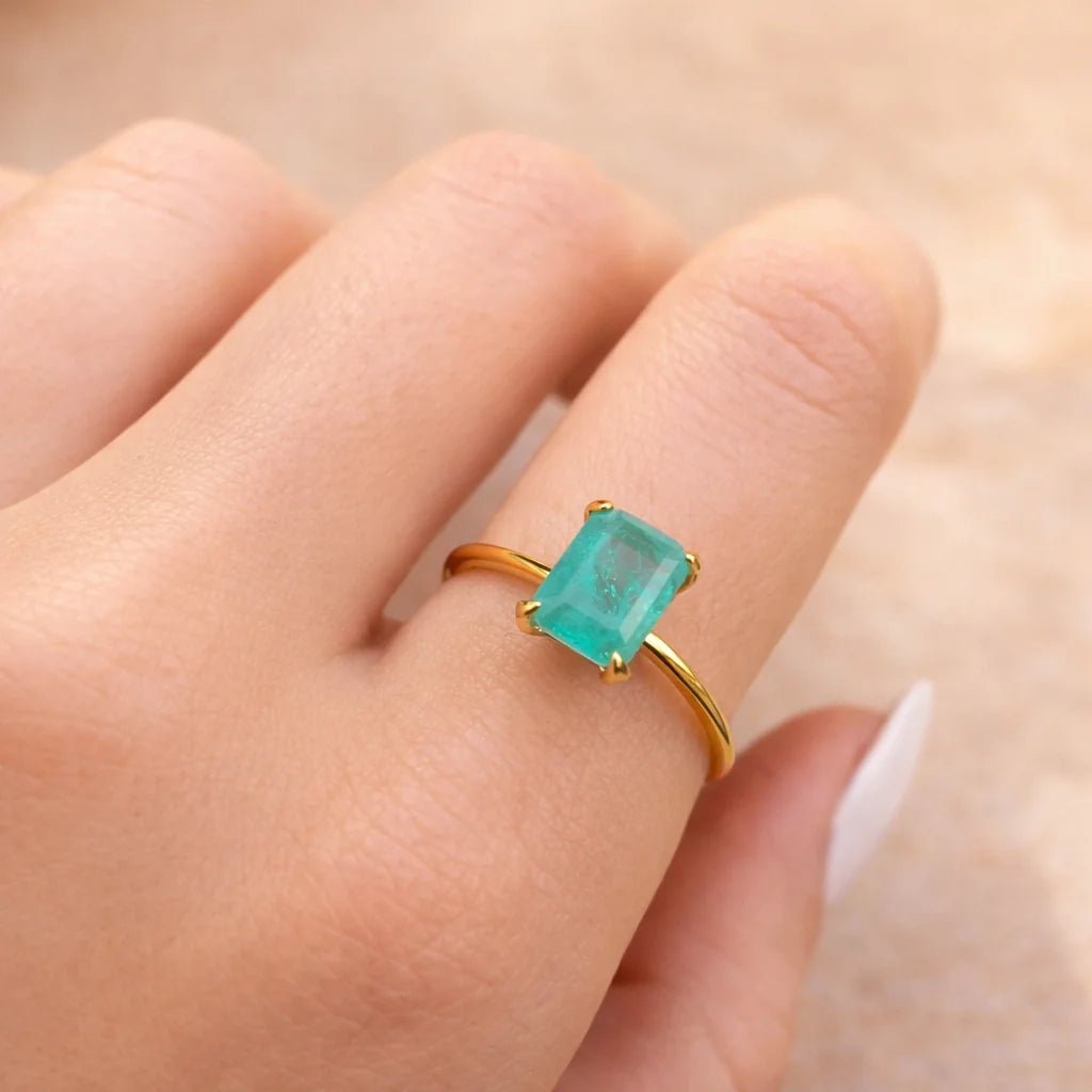 Gold ring with a green blue natural tourmaline gemstone on a finger against a neutral background
