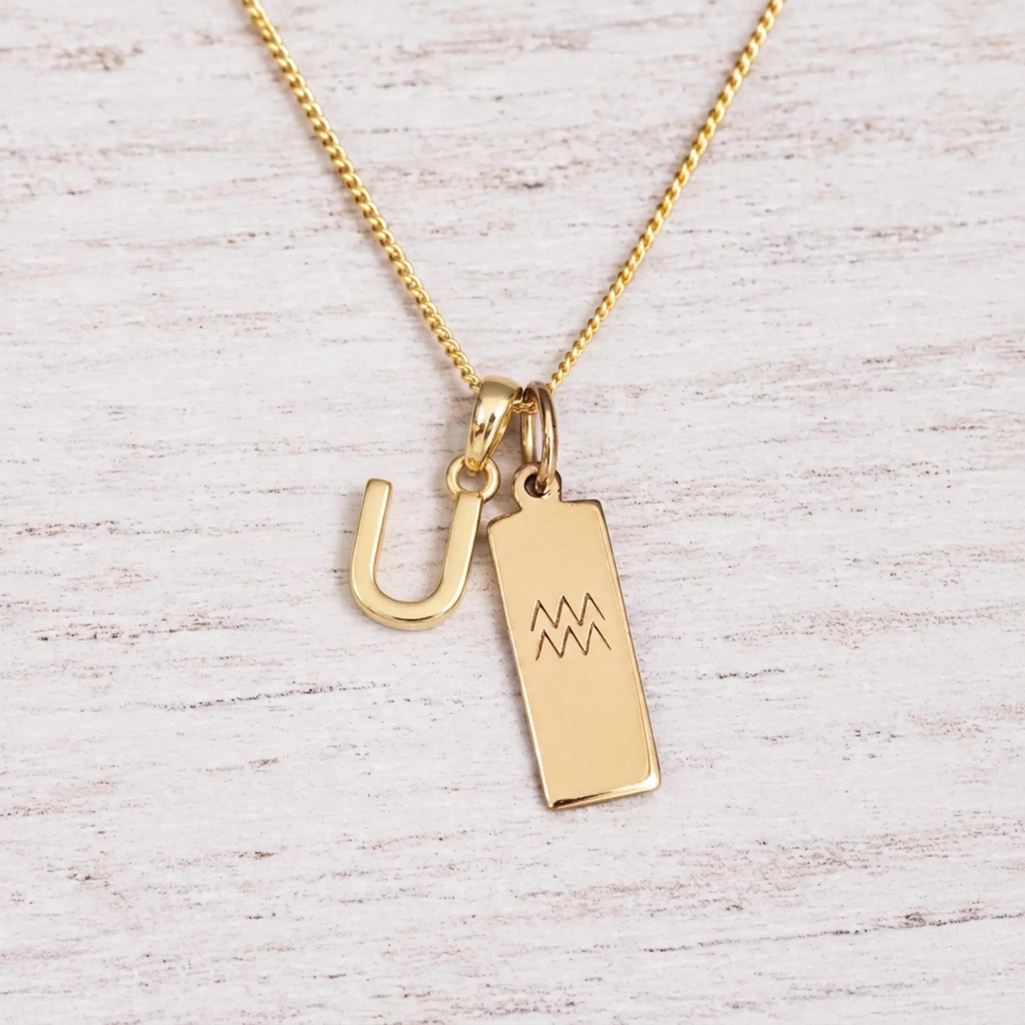 gold U initial and aquarius zodiac necklace on fine gold chain against a light wooden background