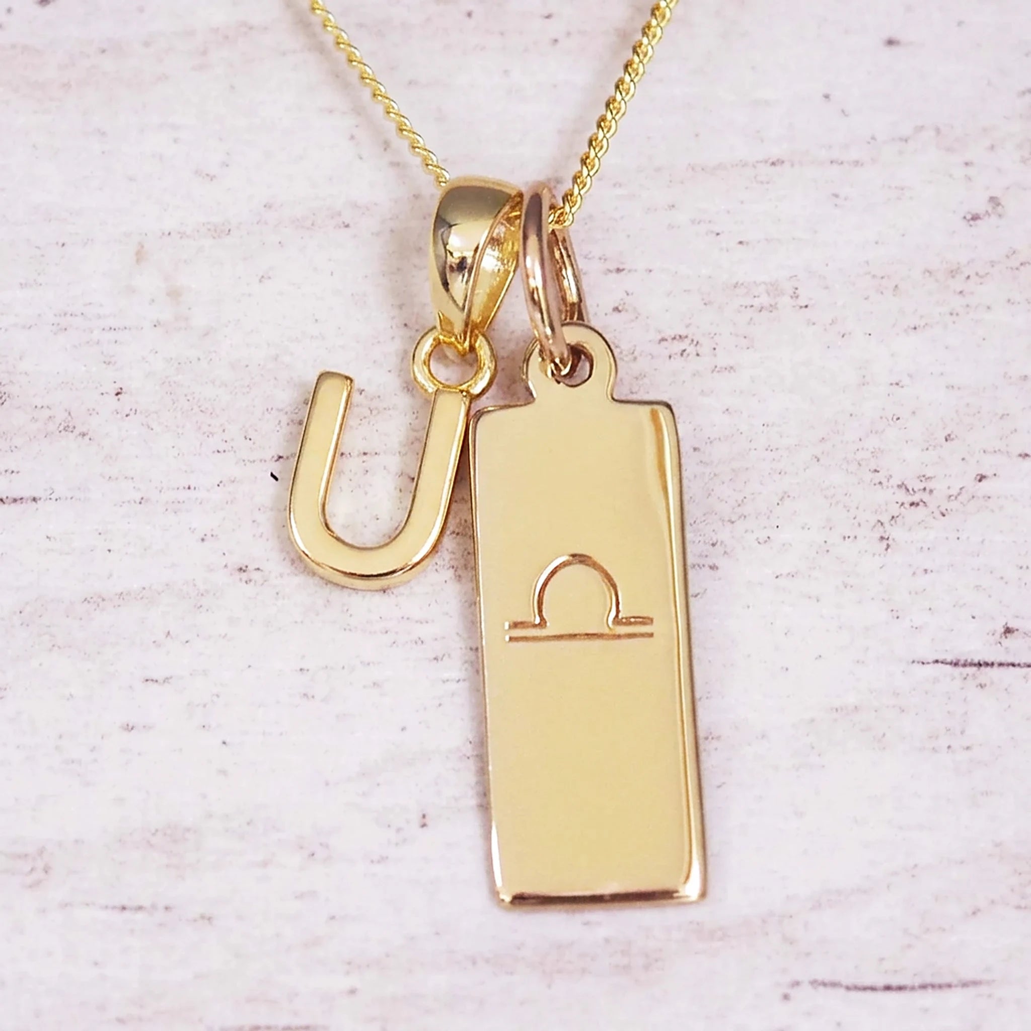 Gold initial and Libra zodiac necklace with a letter U charm and engraved bar pendant on whitewashed wood
