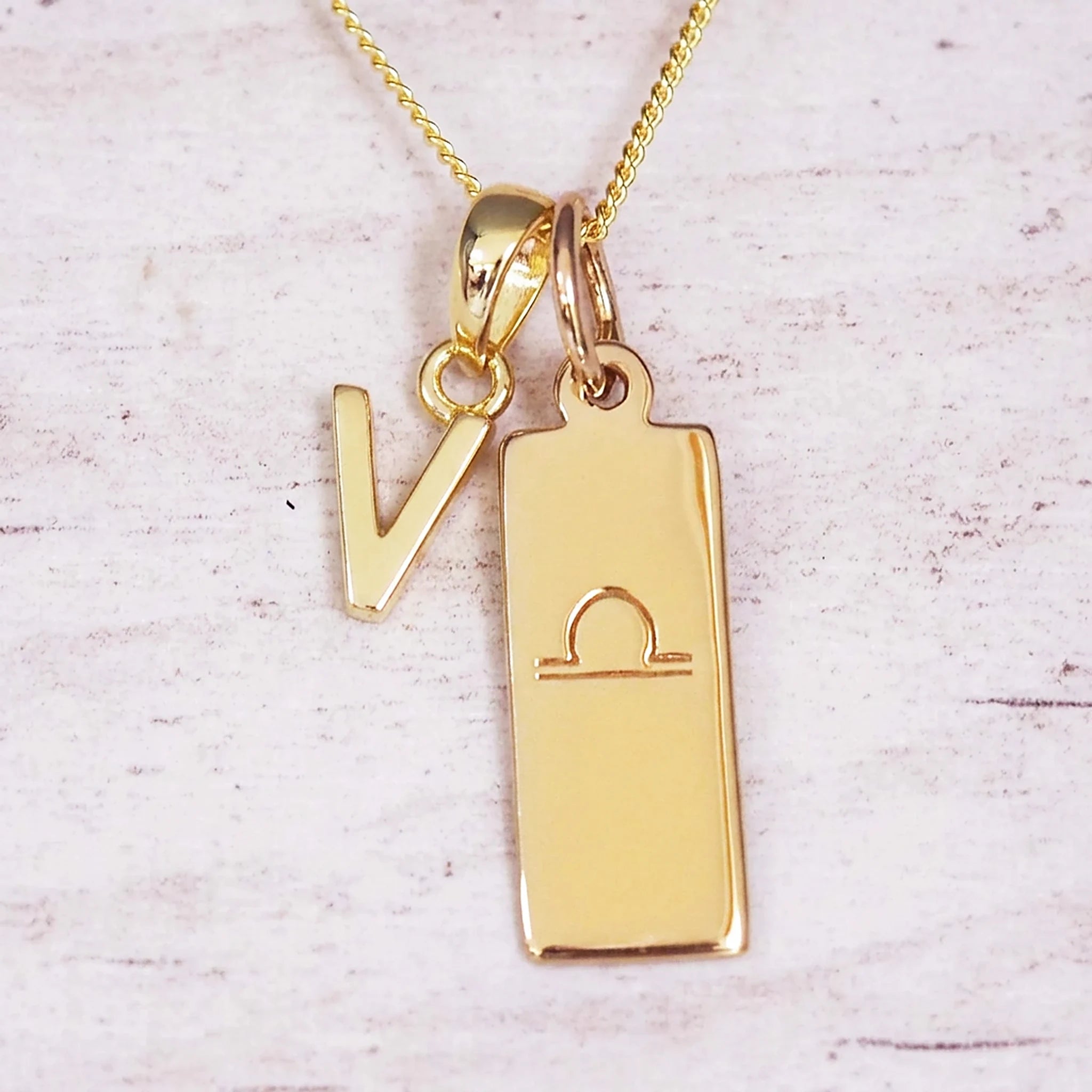 Gold initial and Libra zodiac necklace with a letter V charm and engraved bar pendant on whitewashed wood