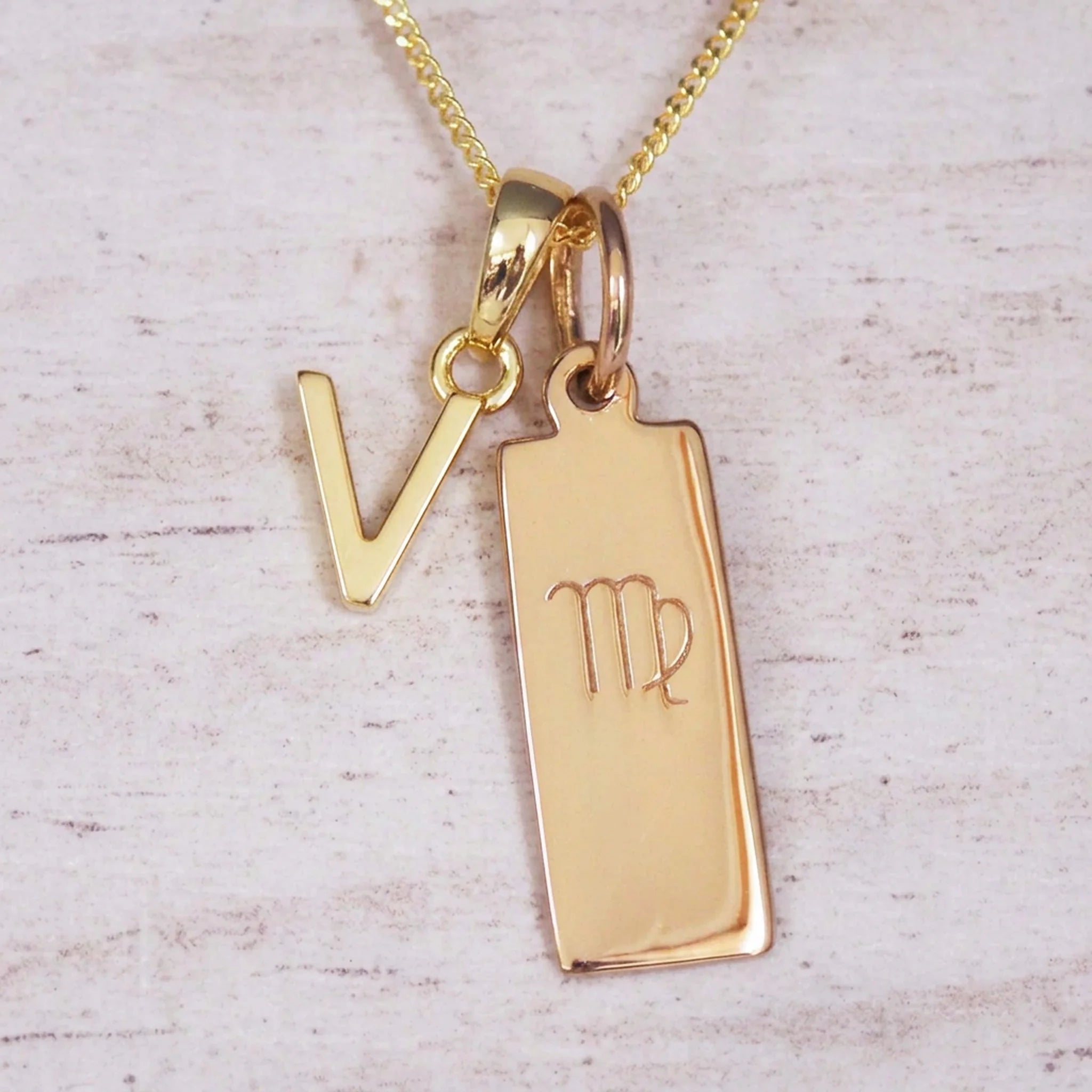 Gold Virgo initial necklace with a letter V pendant and zodiac bar on whitewashed wood