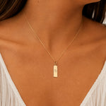 gold virgo zodiac necklace - A close-up of a model's neck and chest wearing a delicate gold chain necklace with a small vertical rectangular pendant engraved with the virgo star sign line glyph.