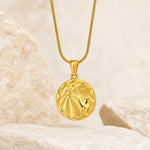 A gold pendant necklace with a circular virgo zodiac sign design, resting on a textured stone with soft dried flowers in a warm, neutral setting.
