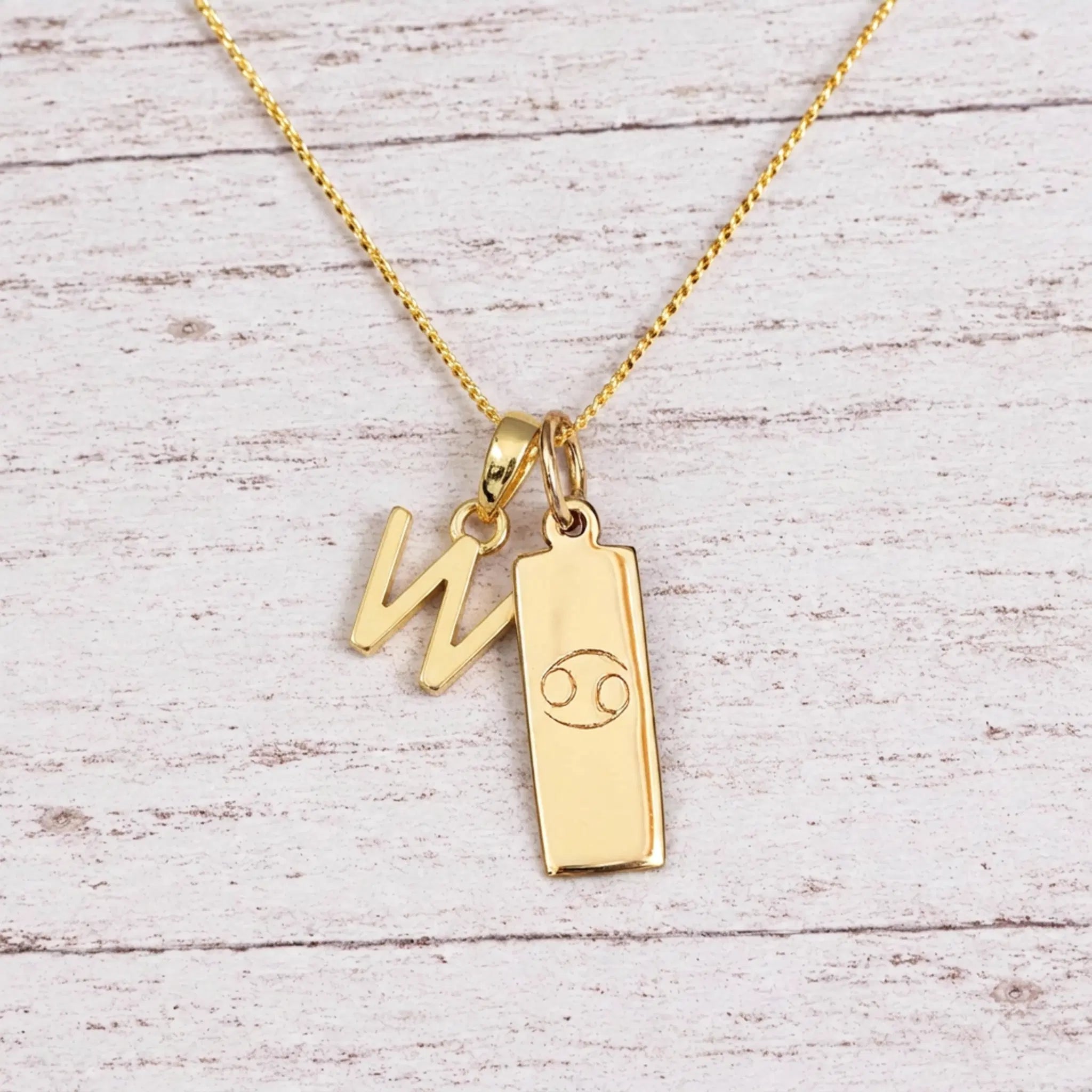 Gold Initial W Pendant and Cancer Zodiac Pendant Necklace on a fine gold chain against a light wooden background
