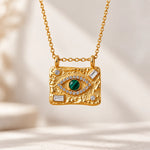 A gold-toned rectangular pendant necklace with a textured finish, featuring a green evil eye motif with clear stones and small rectangular clear stones, hanging from a delicate gold chain against a soft beige background.