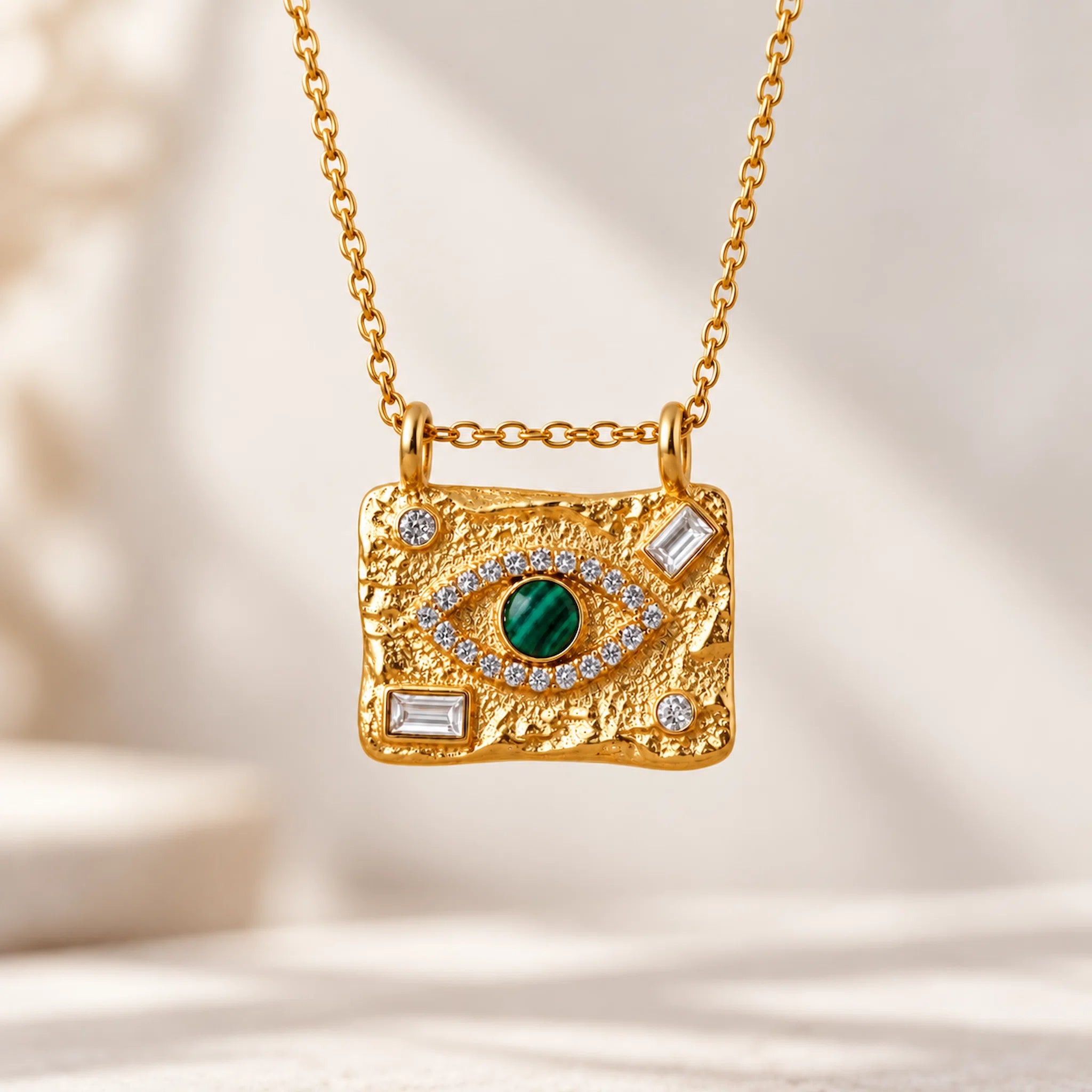 A gold-toned rectangular pendant necklace with a textured finish, featuring a green evil eye motif with clear stones and small rectangular clear stones, hanging from a delicate gold chain against a soft beige background.