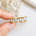 A gold ring with a row of teardrop-shaped white opal gemstones held between two fingers, with dried white flowers in the background.