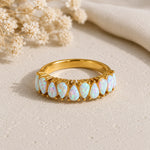 A gold band ring featuring a row of teardrop-shaped white opal gemstones with iridescent blue and pink flashes, displayed on a textured beige surface with dried flowers and a cream fabric in the background.