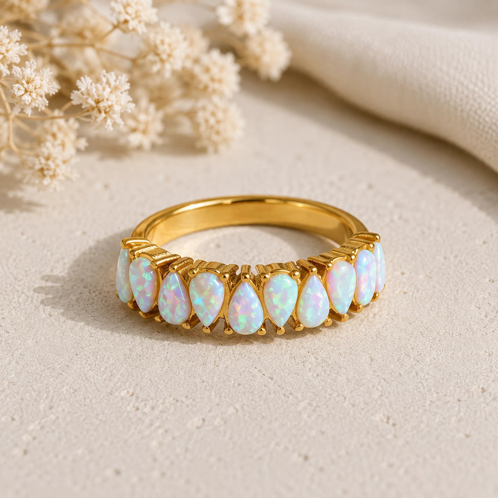A gold band ring featuring a row of teardrop-shaped white opal gemstones with iridescent blue and pink flashes, displayed on a textured beige surface with dried flowers and a cream fabric in the background.