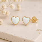 A pair of heart-shaped stud earrings with iridescent white opal inlays and gold borders, displayed on a light beige stone surface with dried flowers in the background. One earring is shown from the front and the other from the side/back, revealing a '925' sterling silver mark on the post.