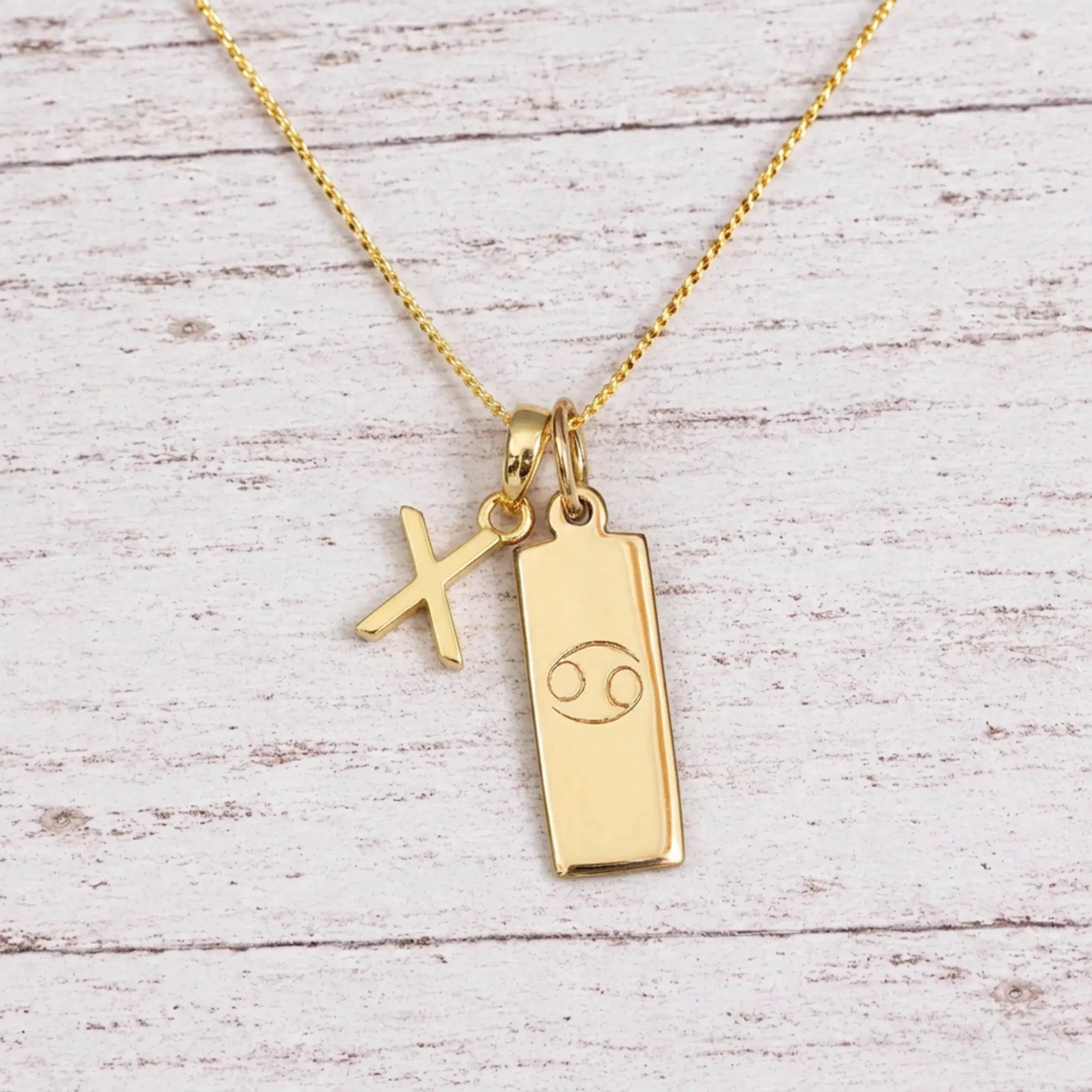 Gold Initial X Pendant and Cancer Zodiac Pendant Necklace on a fine gold chain against a light wooden background