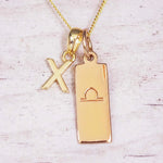 Gold initial and Libra zodiac necklace with a letter X charm and engraved bar pendant on whitewashed wood