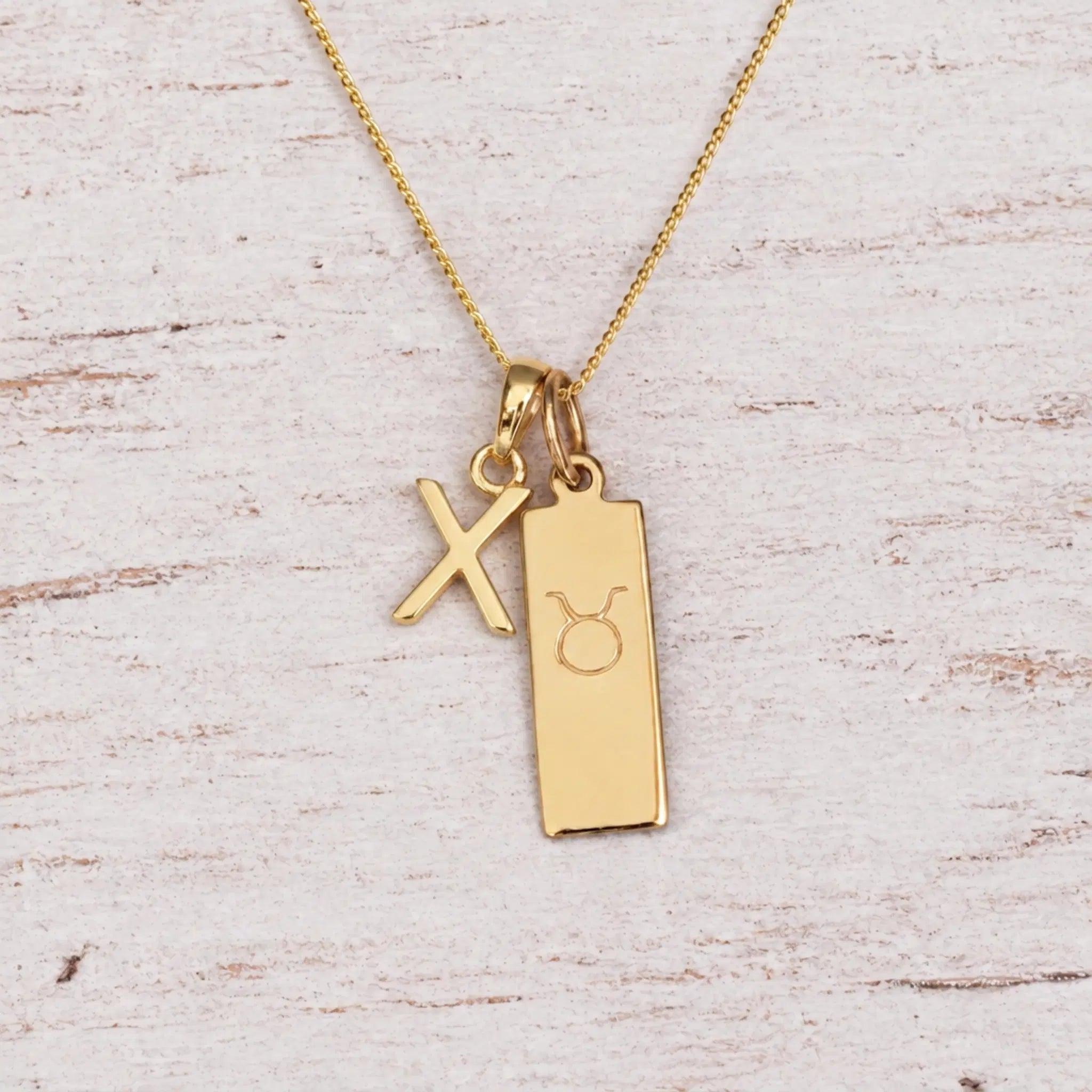 Gold X Initial and Taurus Necklace on a fine gold chain against a light wooden background