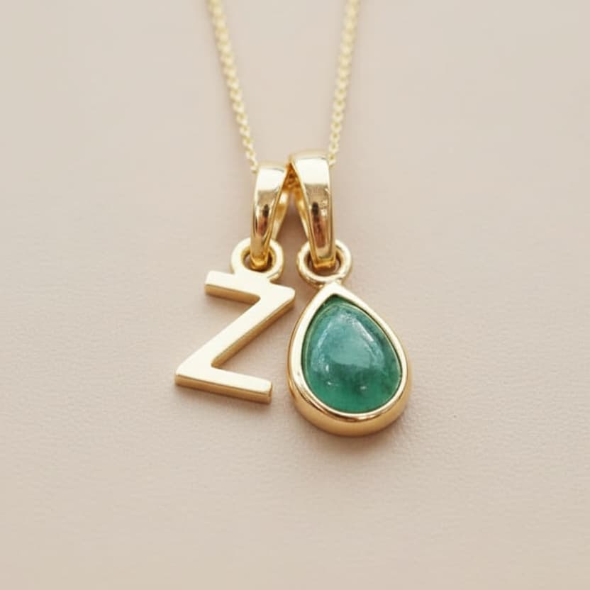 Gold necklace with 'Z' pendant and green natural emerald teardrop pendant on a beige background