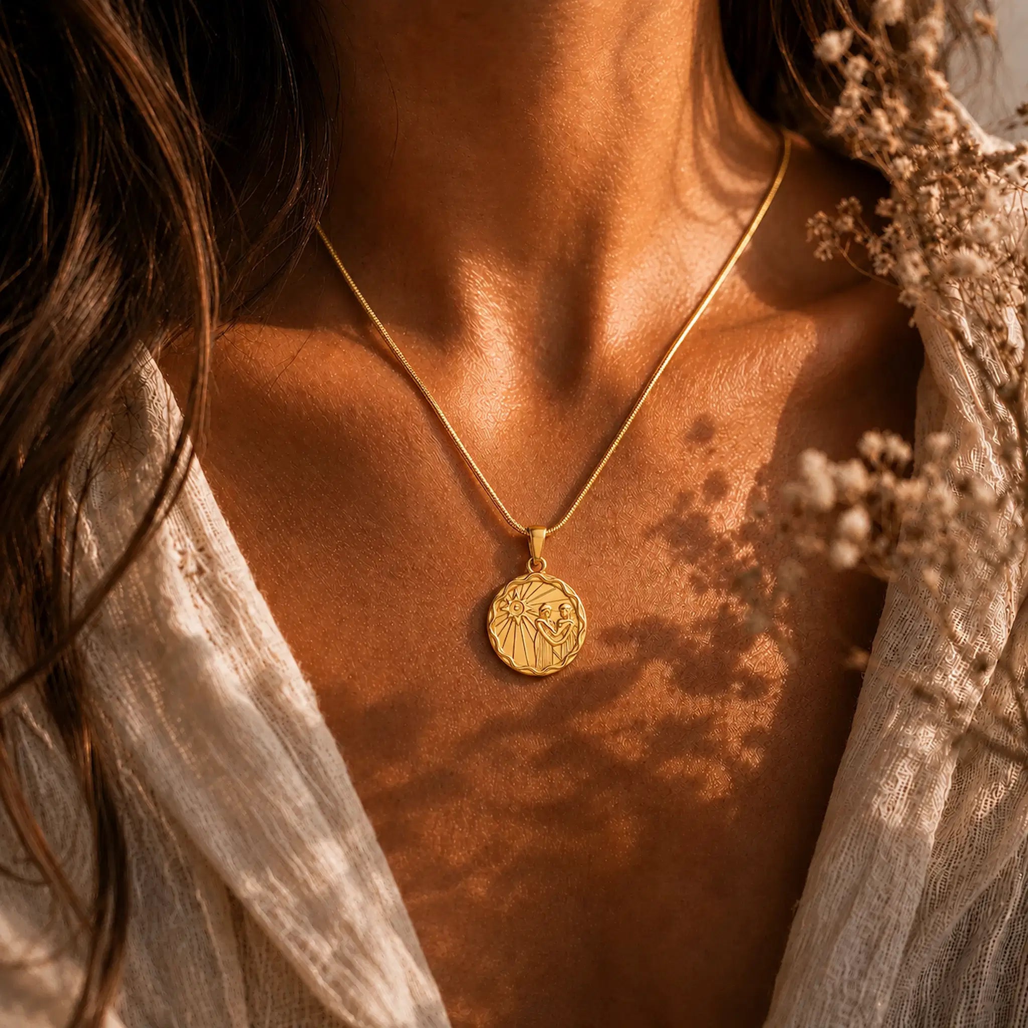 A close-up lifestyle shot of a woman wearing a gold zodiac necklace with a zodiac motif, styled with a white lace top and dried flowers under warm, dappled sunlight.