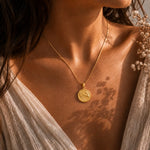 A close-up lifestyle shot of a woman wearing a gold coin pendant necklace. The pendant features an embossed zodiac design, hanging from a delicate gold chain.
