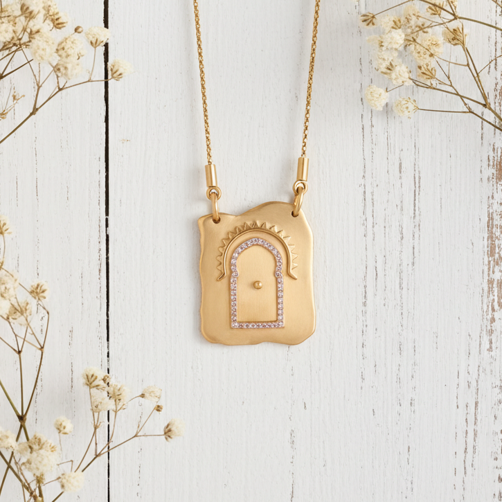 Gold necklace with a decorative pendant on a white wooden surface with dried flowers.