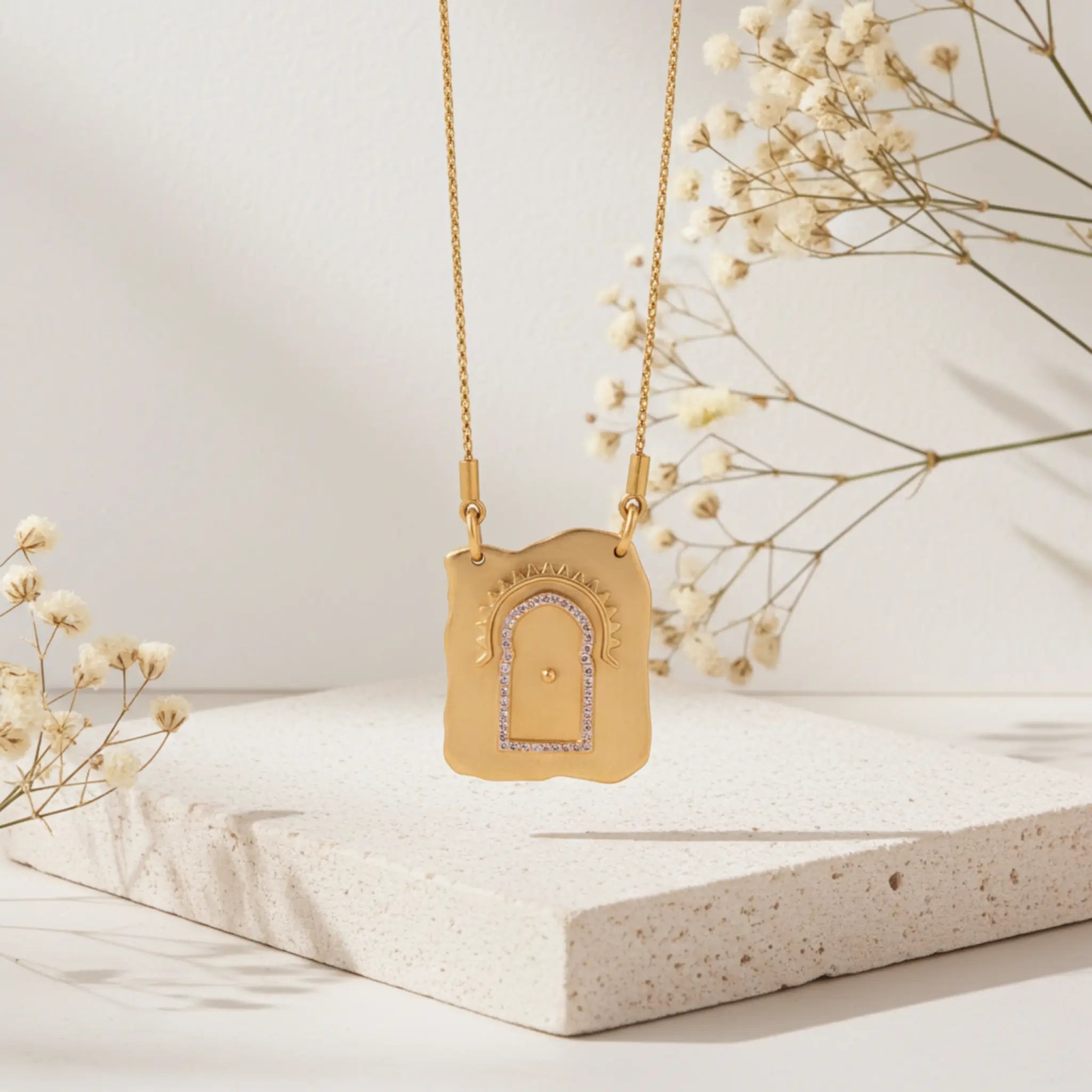 Gold necklace with a decorative pendant on a light surface with dried flowers.