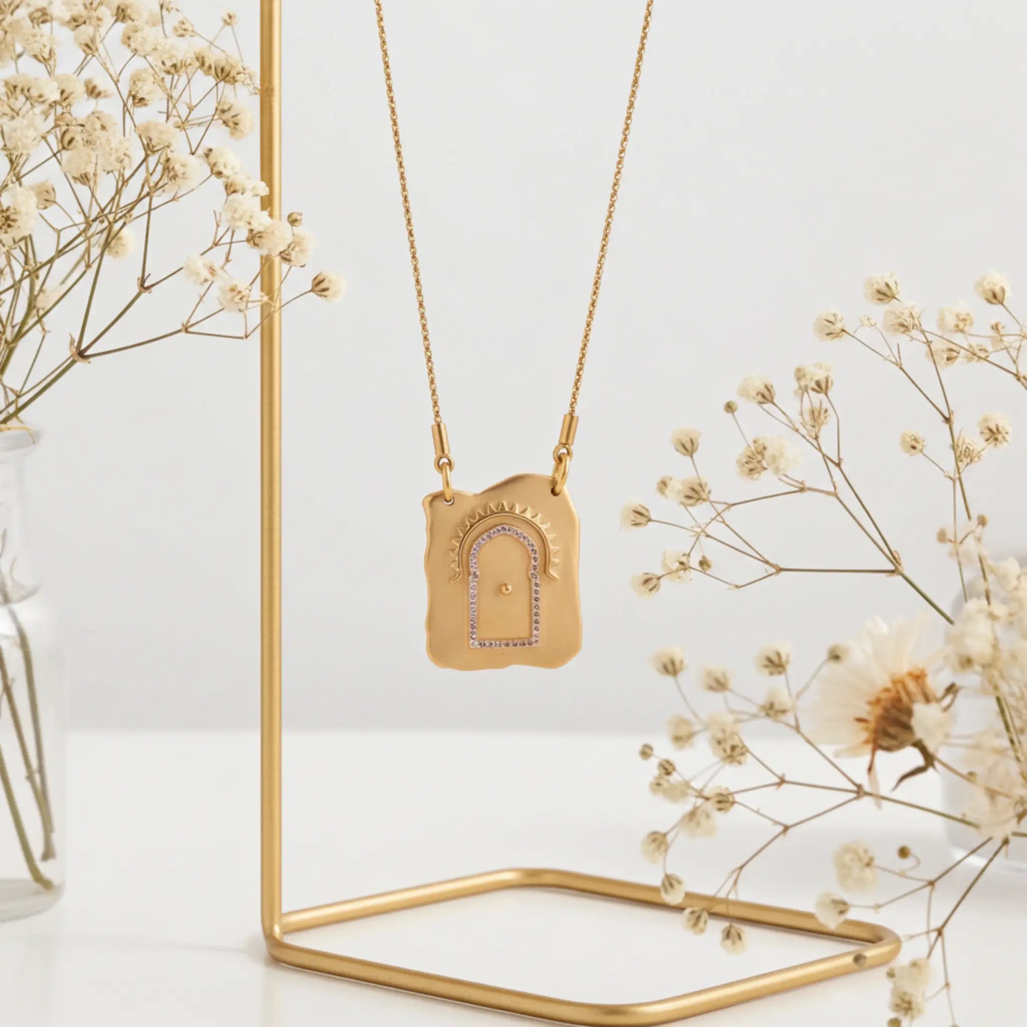 Gold necklace with a pendant on a gold stand, surrounded by dried flowers.