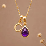 Gold initial pendant necklace featuring the letter 'C' and a february birthstone teardrop-shaped natural amethyst gemstone charm, displayed against a warm beige background with subtle dried flowers.