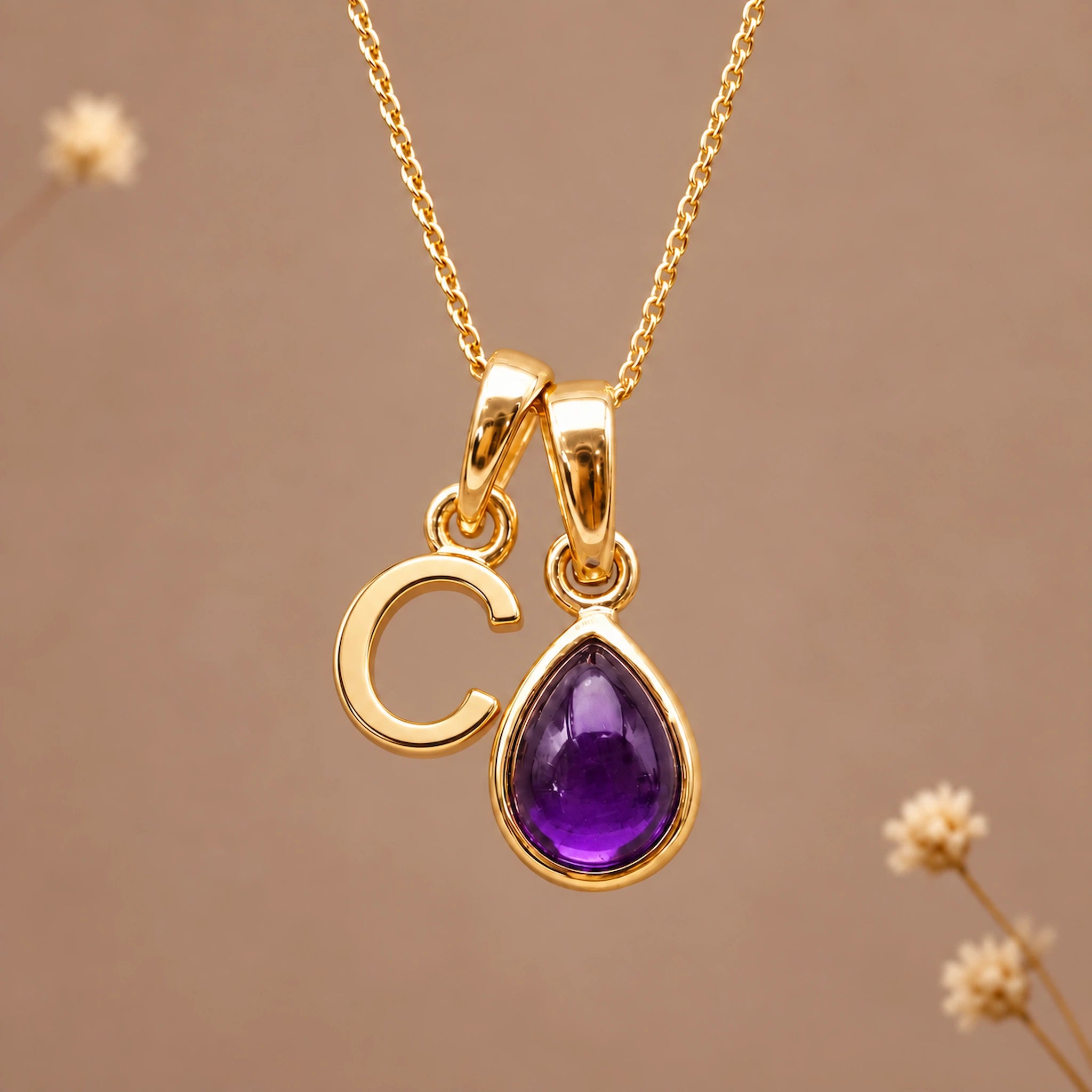 Gold initial pendant necklace featuring the letter 'C' and a february birthstone teardrop-shaped natural amethyst gemstone charm, displayed against a warm beige background with subtle dried flowers.