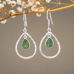 Silver earrings with green teardrop stones on a branch with small white flowers.