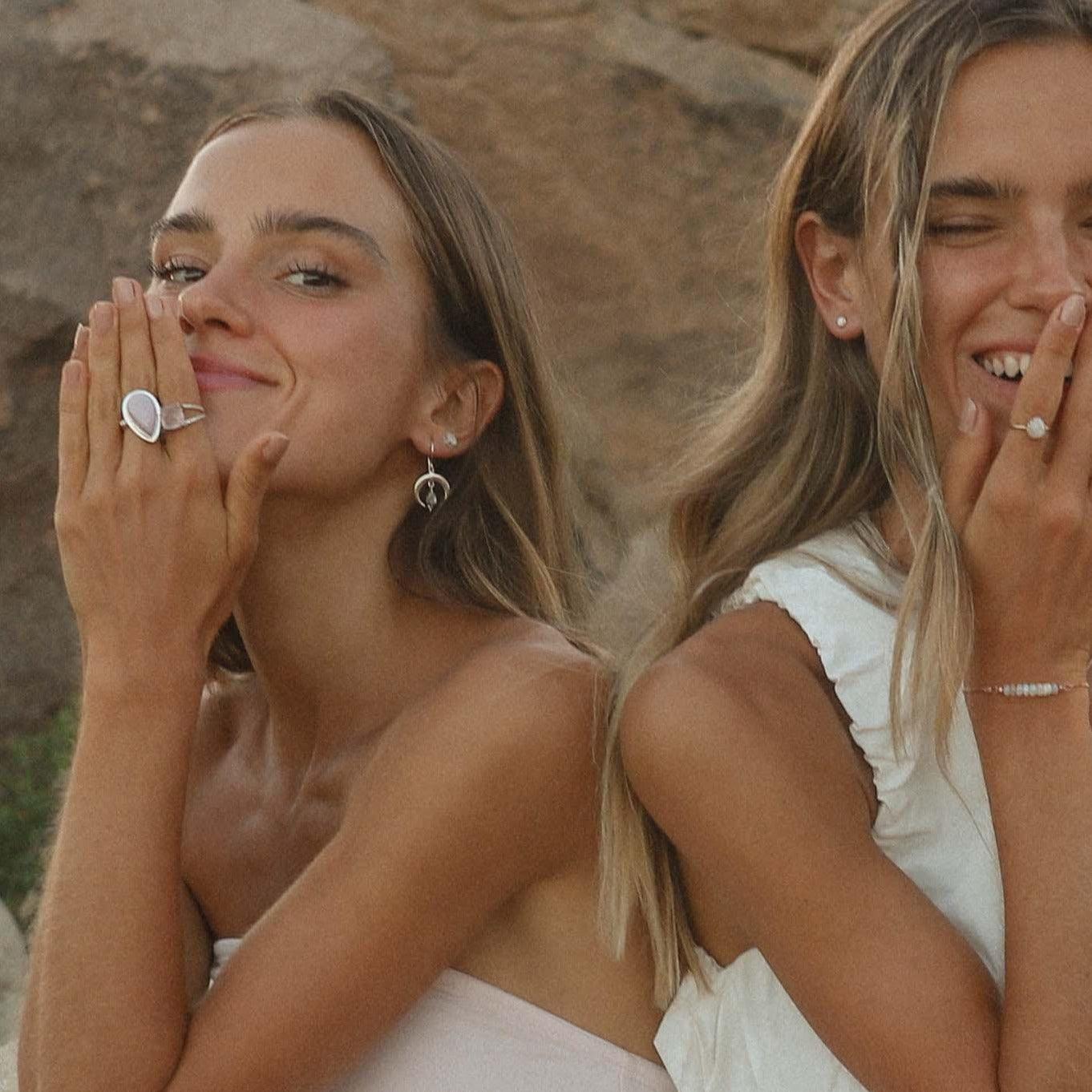 Two women with blonde hair and earrings, one wearing a ring, against a natural stone background.