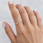 A close-up of a hand wearing a thin, hammered textured sterling silver stacker band ring, set against a soft, light gray background.