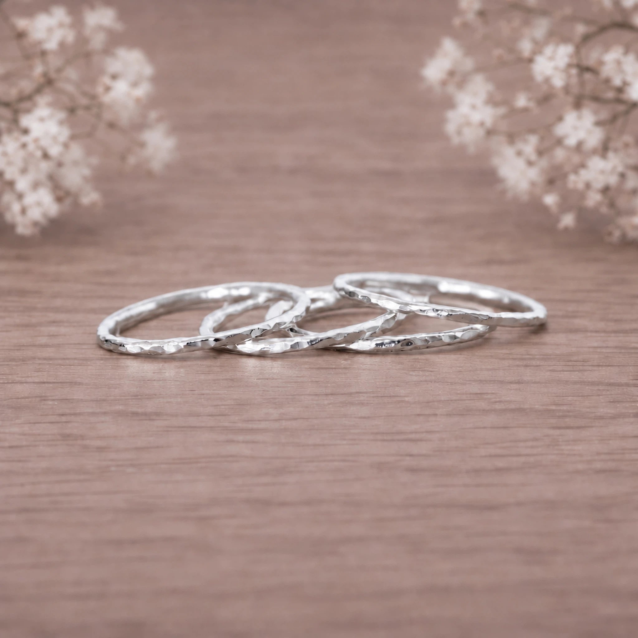 four silver rings with a hammered texture, stacked together on a wooden surface with soft-focus baby's breath flowers in the background.