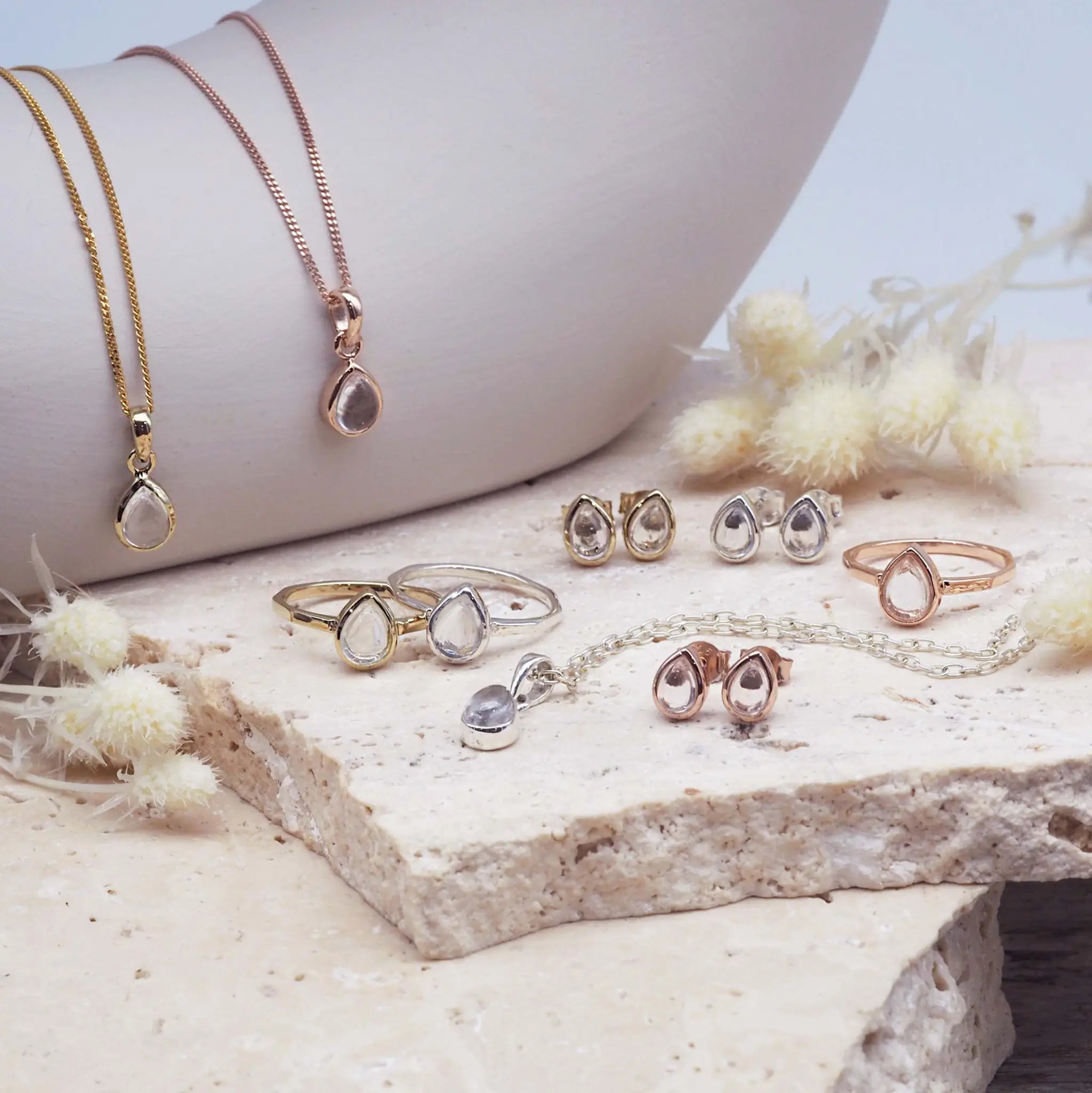 Collection of silver, gold and rose gold april birthstone herkimer quartz jewellery including necklaces, rings, and earrings on a stone surface with dried flowers. indie and harper jewellery australia