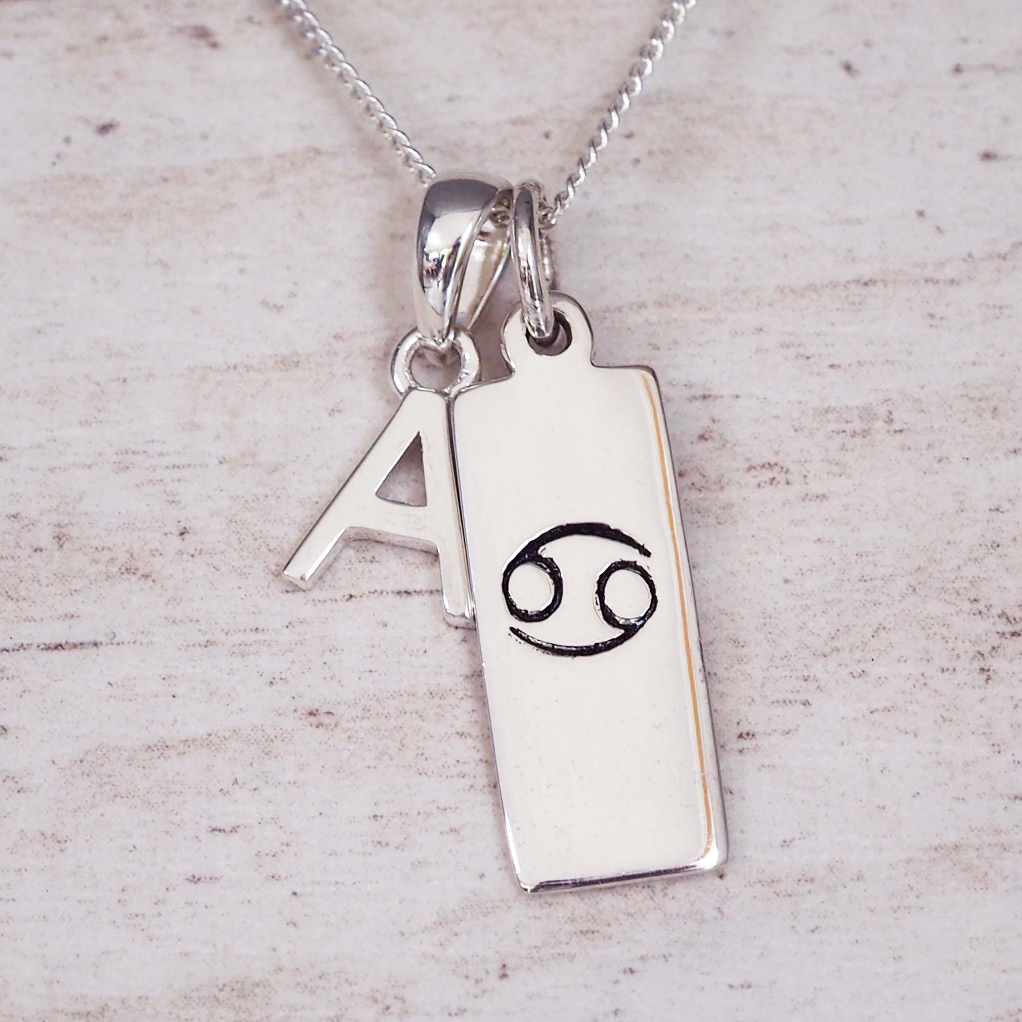 Silver necklace with a pendant featuring the letter 'A' and a Cancer zodiac symbol on a light wooden background.