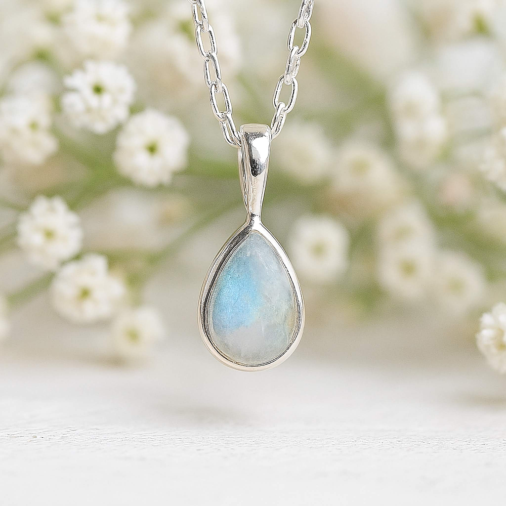 Teardrop-shaped moonstone pendant with a blue-green gemstone on a sterling silver chain, set against a blurred background of white flowers.