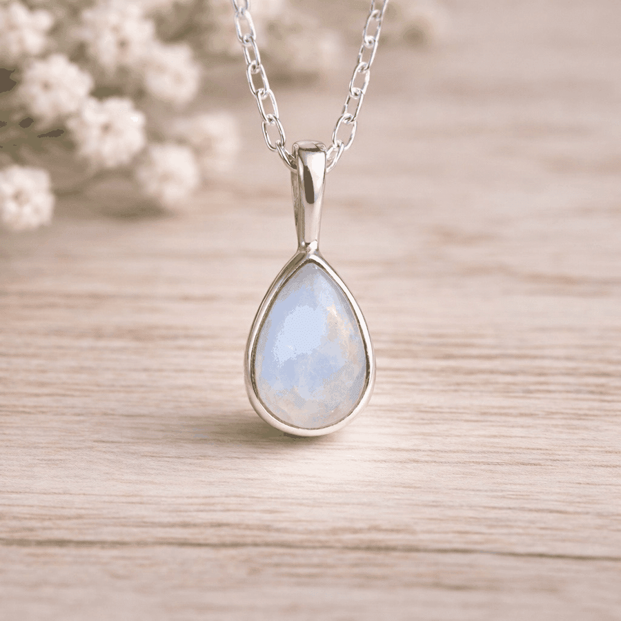 Teardrop-shaped moonstone pendant with a blue-green gemstone on a sterling silver chain, set against a blurred background of white flowers.