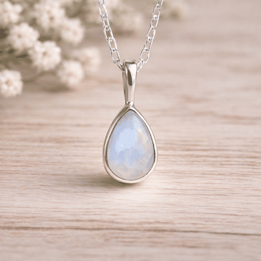 Teardrop-shaped moonstone pendant with a blue-green gemstone on a sterling silver chain, set against a blurred background of white flowers.
