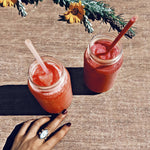 Two jars of red smoothie with straws on a wooden surface, hand with ring visible.