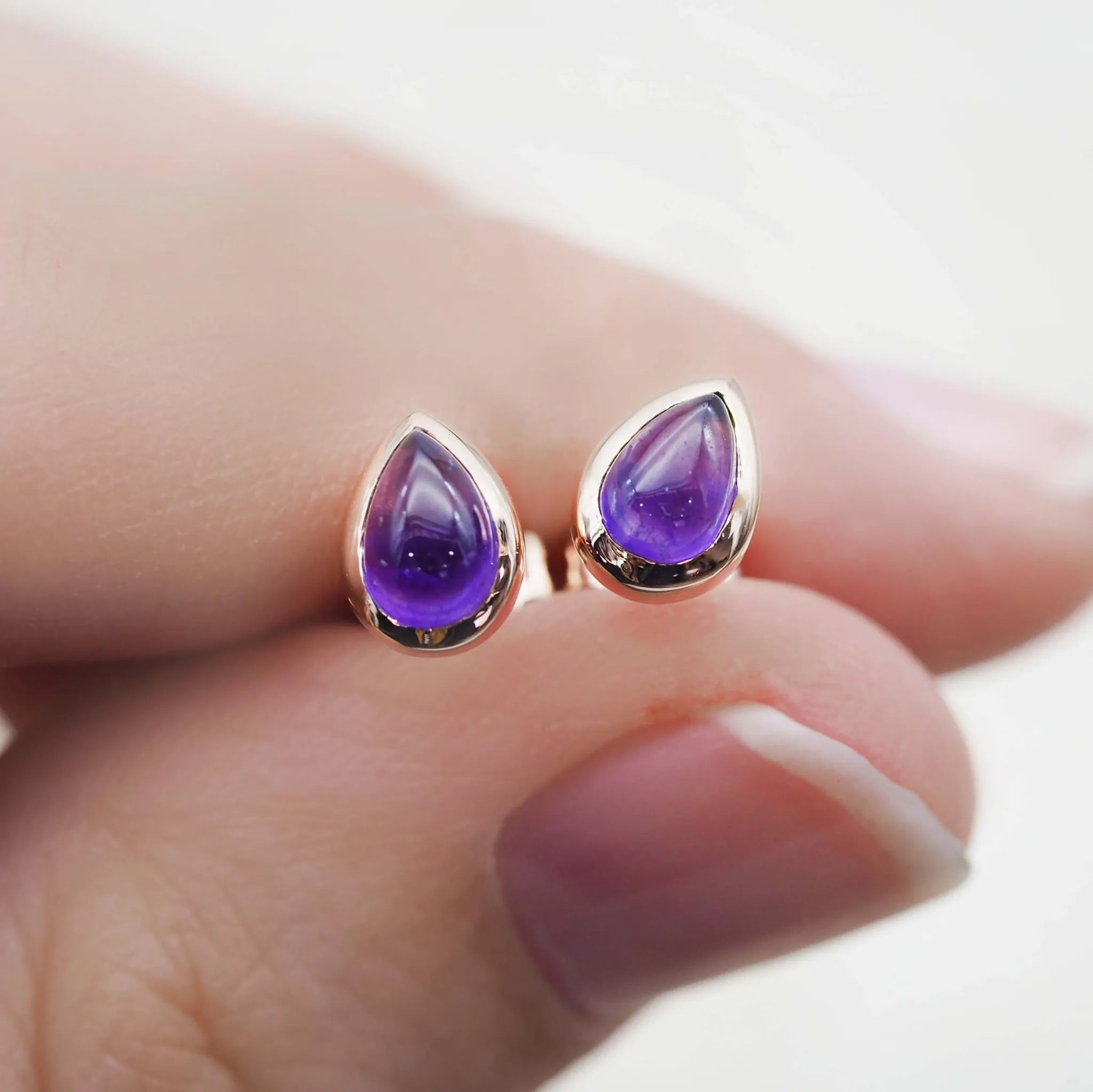 Teardrop-shaped purple gemstone amethyst earrings in rose gold held between fingers on a light background - indie and harper 