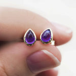 Purple teardrop earrings with gold accents held between fingers against a blurred background